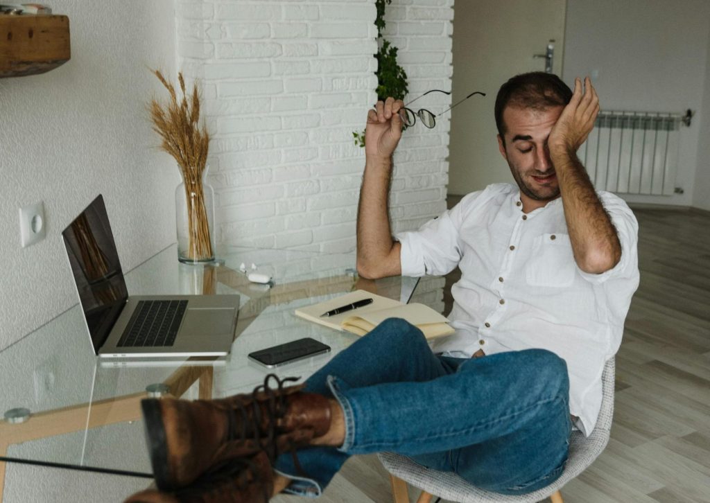 Man in casual attire sitting in a home office with his feet up, appearing tired while working remotely.