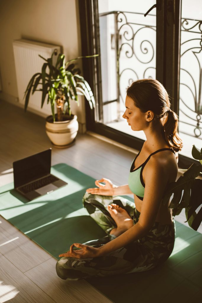 A woman meditates in lotus pose on a yoga mat at home, guided by a laptop.